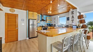 Eine moderne Küche mit Holzausstattung, einer zentralen Kochinsel mit Barhocker, einem Edelstahlkühlschrank und Blick ins Freie.