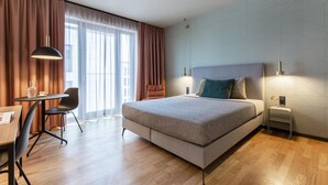 Une chambre d’hôtel moderne, dotée d’un grand lit, d’un bureau avec une chaise et offrant une vue sur la ville grâce à de grandes fenêtres.