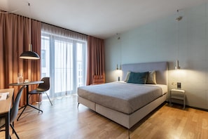 Ein modernes Hotelzimmer mit einem großen Bett, einem Schreibtisch mit Stuhl und Blick auf die Stadt durch große Fenster.