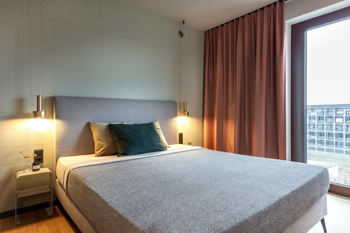 Ein modernes Hotelzimmer mit einem großen Bett, zwei Nachttischlampen, einem Nachttisch und Blick durch ein großes Fenster.