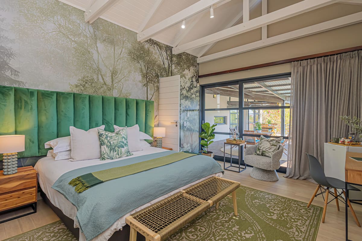 Een moderne slaapkamer met een groot bed, een groen hoofdeinde, een bankje en uitzicht op het omliggende landschap door een glazen deur.