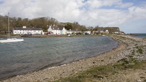 A coastal town with a row of white buildings along the shore, a boat docked near the water, and a rocky beach. - 2 The Coach House (Pentraeth)