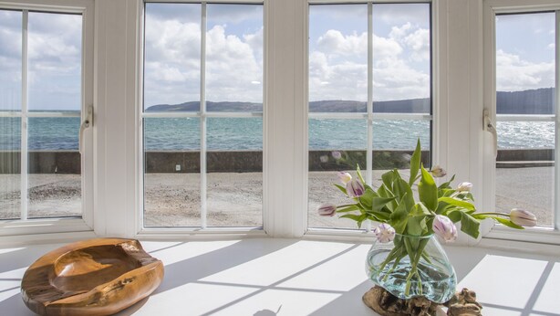 A room with large windows overlooking a beach and sea, a wooden bowl, and a vase with flowers. - 2 The Coach House (Pentraeth)