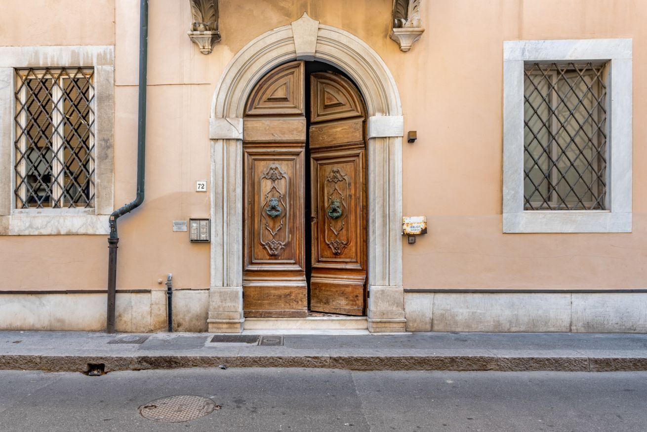 Un edificio con una puerta doble de madera, un ventanal pequeño con celosía metálica y una placa con un número.