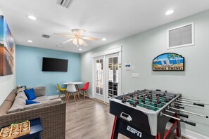 A room with a sofa, a table with chairs, a foosball table, a television, and a wall-mounted sign. - Just Beachy Private Pool! Just 20 Steps to the beach! Incredible Home (PANAMA CITY BEACH)