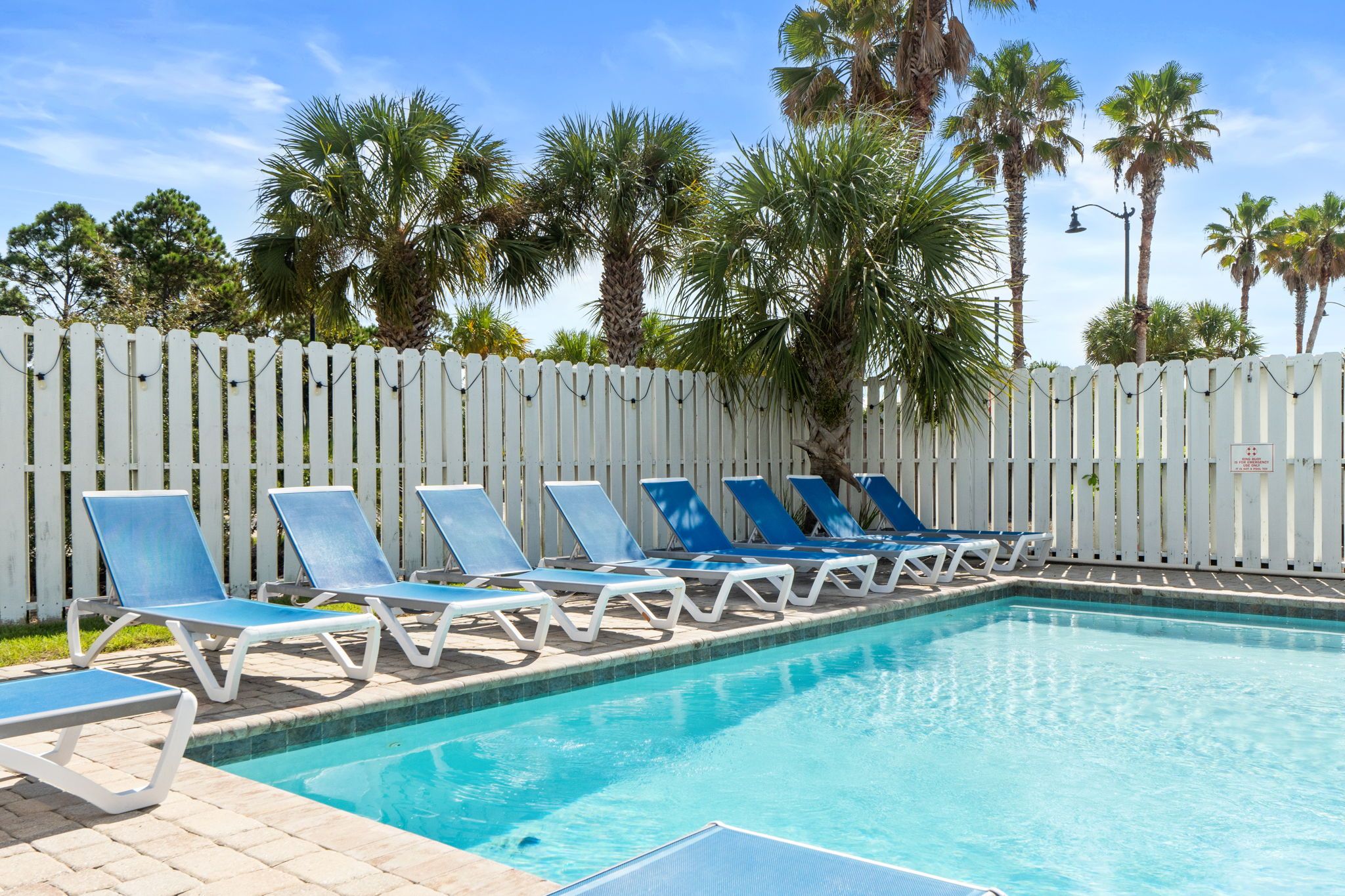 A swimming pool with blue lounge chairs and a white fence in the background.