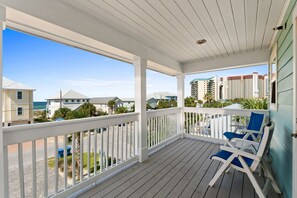 A covered porch with white railings, two blue lounge chairs, and a view of a beach and buildings in the background. - Just Beachy Private Pool! Just 20 Steps to the beach! Incredible Home (PANAMA CITY BEACH)