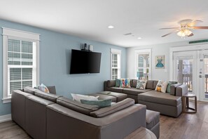 A modern living room with a sectional sofa, a flat-screen TV, and a ceiling fan. - Just Beachy Private Pool! Just 20 Steps to the beach! Incredible Home (PANAMA CITY BEACH)