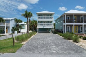 A two-story beachfront house with a paved driveway, palm trees, and a clear sky.
