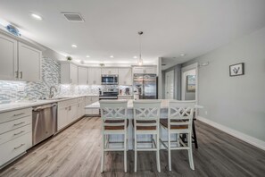 A modern kitchen with white cabinets, a tiled backsplash, stainless steel appliances, and a bar area with white bar stools.