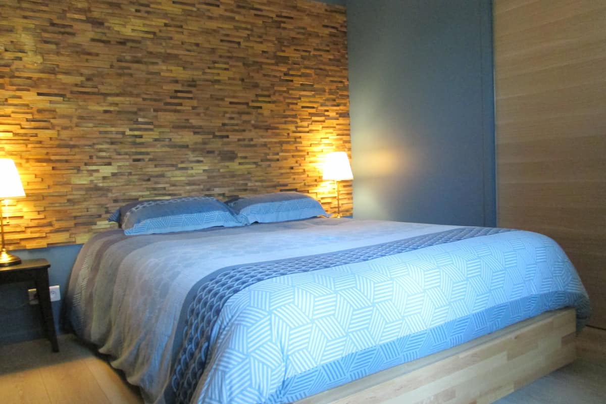A bedroom with a wooden headboard, a patterned bedspread, two bedside lamps, and a brick-patterned wall.