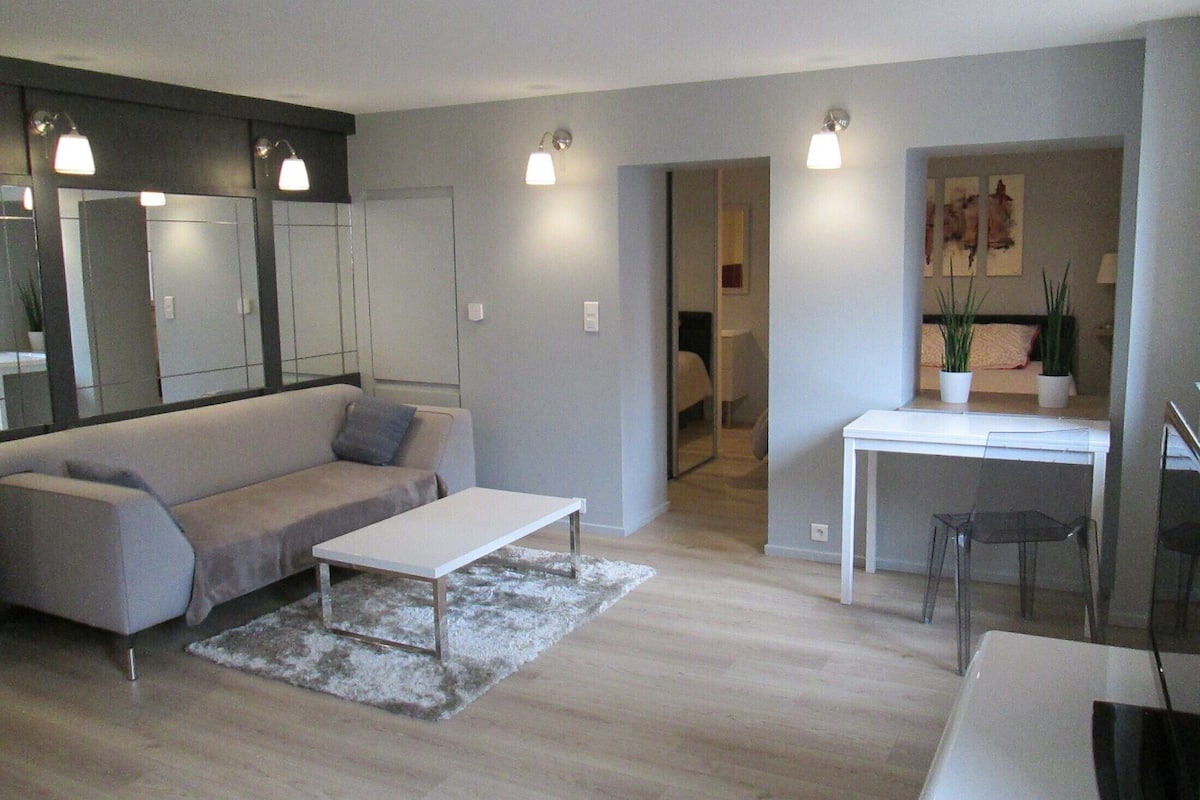A modern living room with a grey sofa, a white coffee table, and a small rug. There is a desk with a chair and a potted plant.