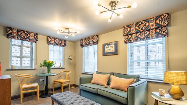 A cozy living room with a green sofa, a wooden bench, and a small table with a lamp. There are two windows with patterned curtains, a framed picture on the wall, and a vase with flowers on a table. - The Falcon - Apartment 1 (York)