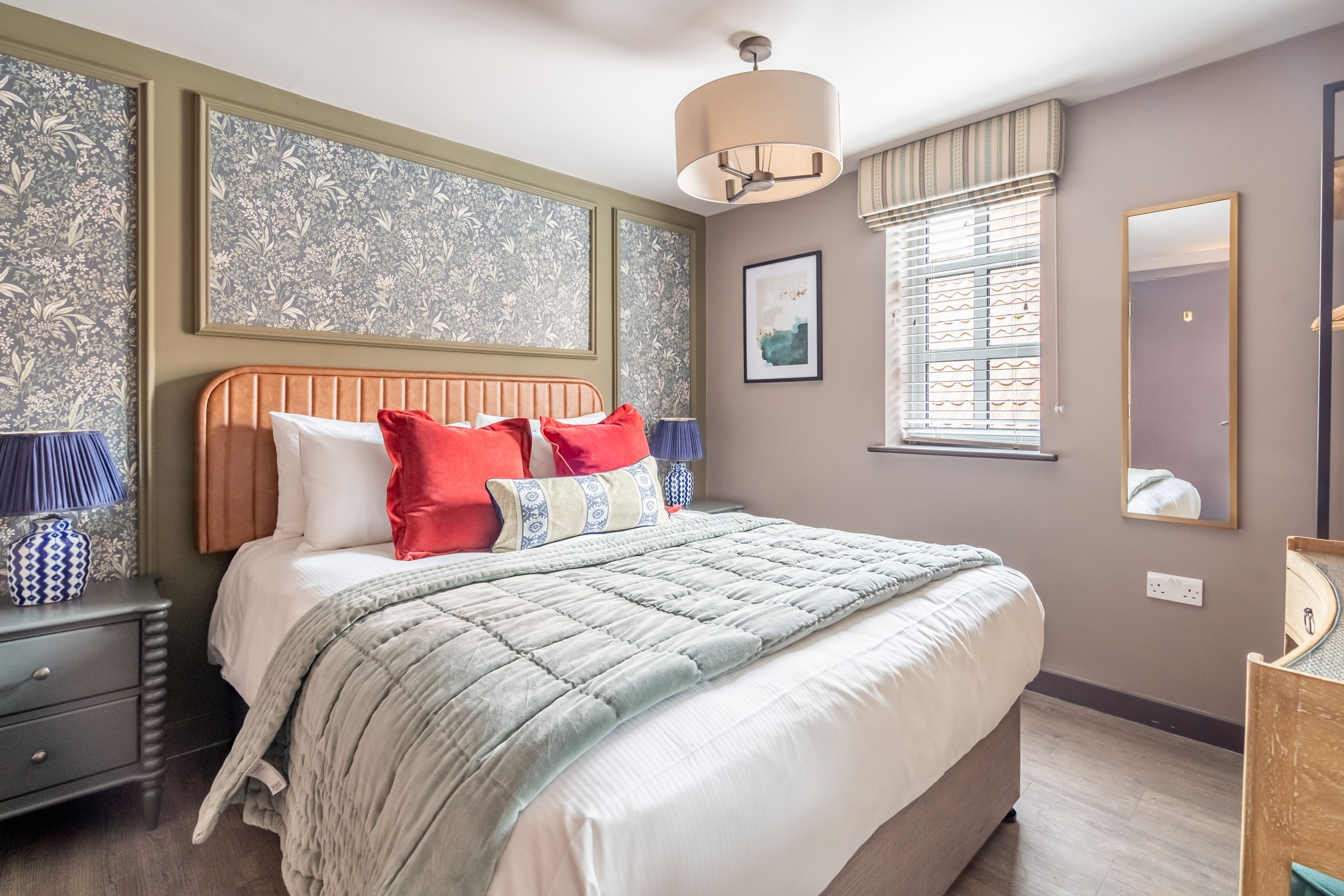 A bedroom with a large bed, a nightstand, a lamp, a mirror, and a window with blinds.
