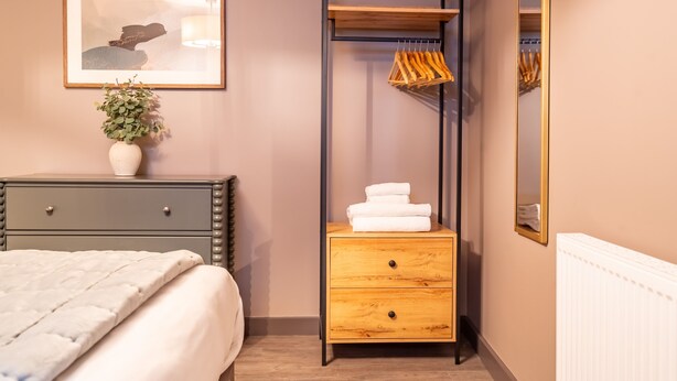 A bedroom with a bed, a bedside table with a vase, a wooden dresser with drawers, a hanging rack, and a mirror. - The Falcon Apartment 5 (York)