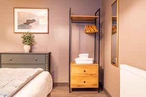 A bedroom with a bed, a bedside table with a vase, a wooden dresser with drawers, a hanging rack, and a mirror. - The Falcon Apartment 5 (York)