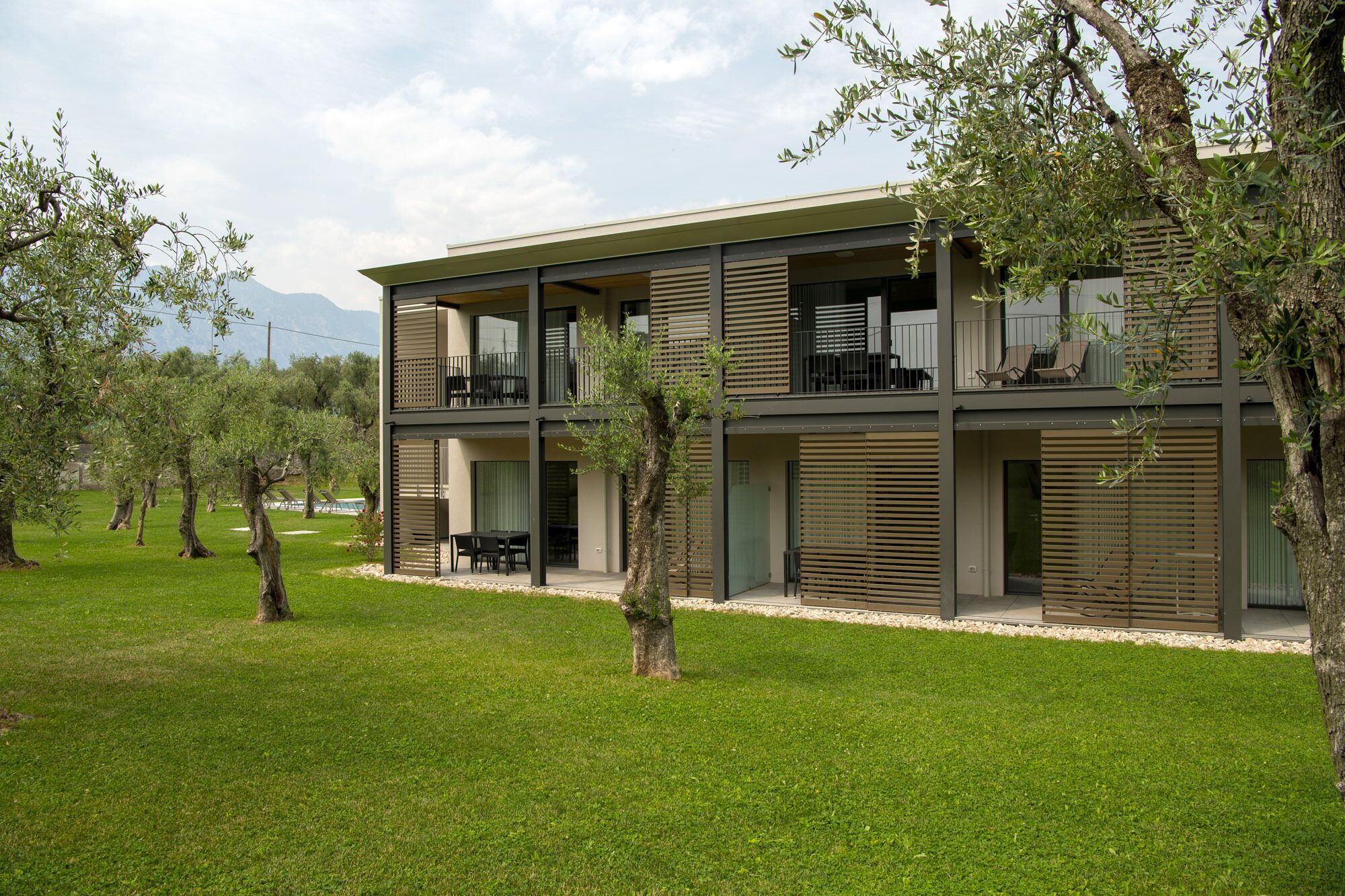 Un bâtiment moderne à deux étages, doté de balcons, entouré d’une pelouse bien entretenue et d’oliviers.