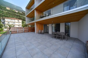 A modern two-story building with a wooden balcony, a dining area with chairs and a table, and a view of the surrounding hillside.