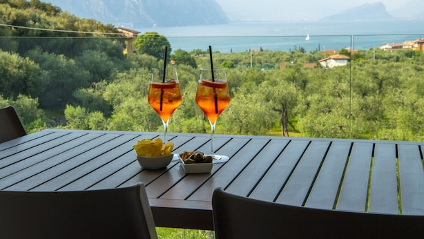Una mesa con dos vasos de una bebida, un bol de patatas fritas y un plato con aperitivos. La mesa está al aire libre, con vistas a un paisaje montañoso y una masa de agua al fondo.