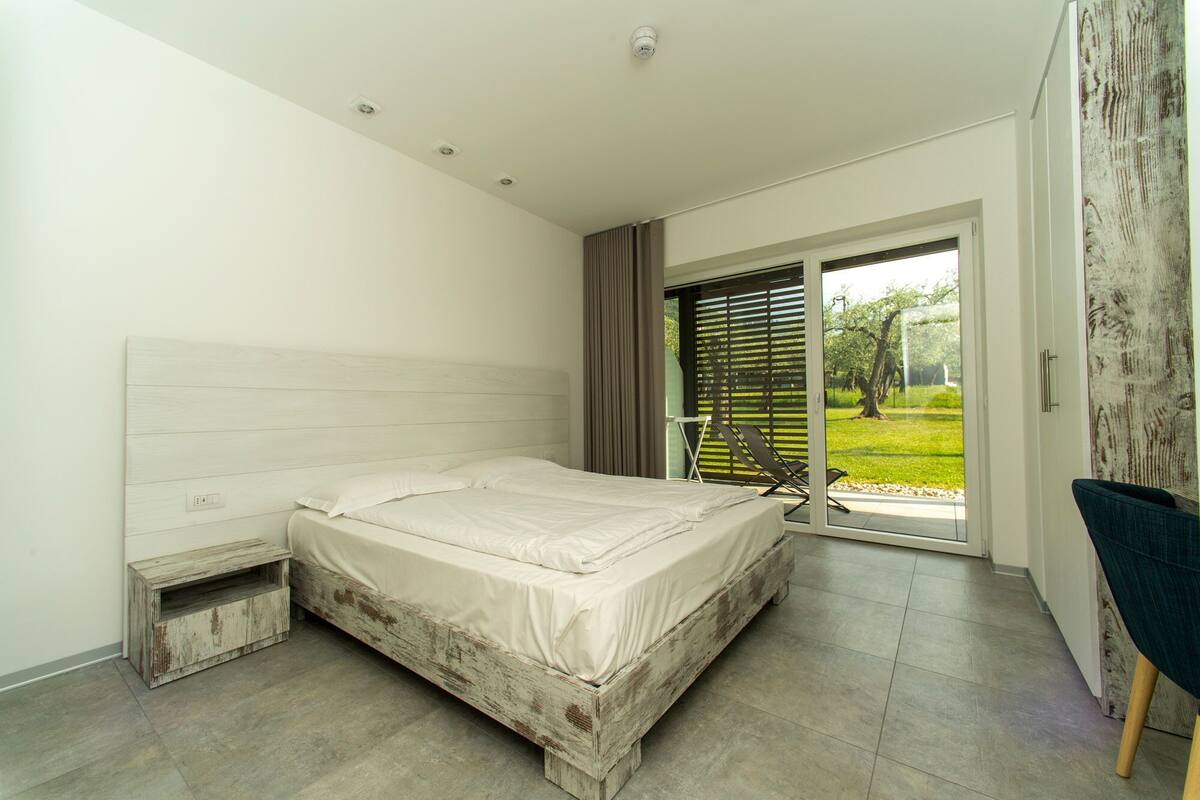 Un dormitorio con cama de madera, una mesita de noche pequeña, una silla y vistas a un jardín a través de una puerta corredera.