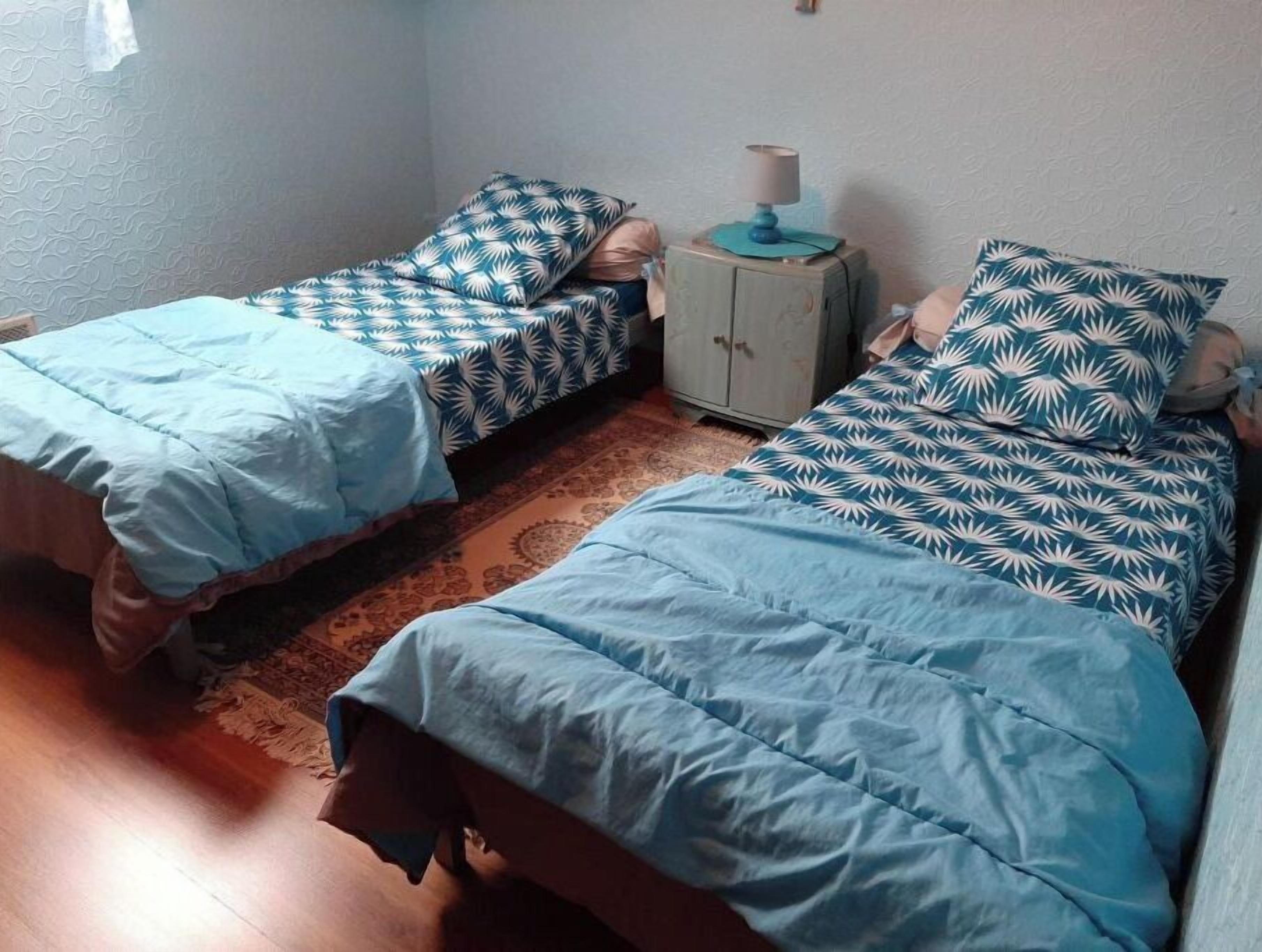 A room with two single beds, each with a patterned duvet cover, a small bedside table with a lamp, and a patterned rug on the floor.