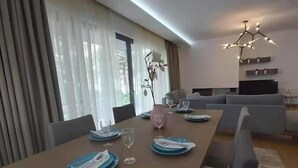 A modern dining area with a long table set for four, a sofa, and a TV in the background. - GTD35501 - Unique Villa in Voula Riviera (Vari-Voula-Vouliagmeni)