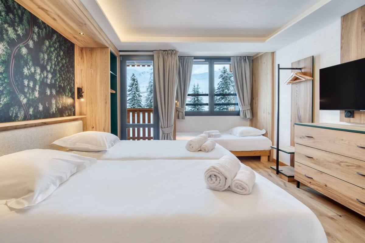 Habitación de hotel con dos camas, televisor, cómoda de madera y balcón con vista a árboles y montañas.