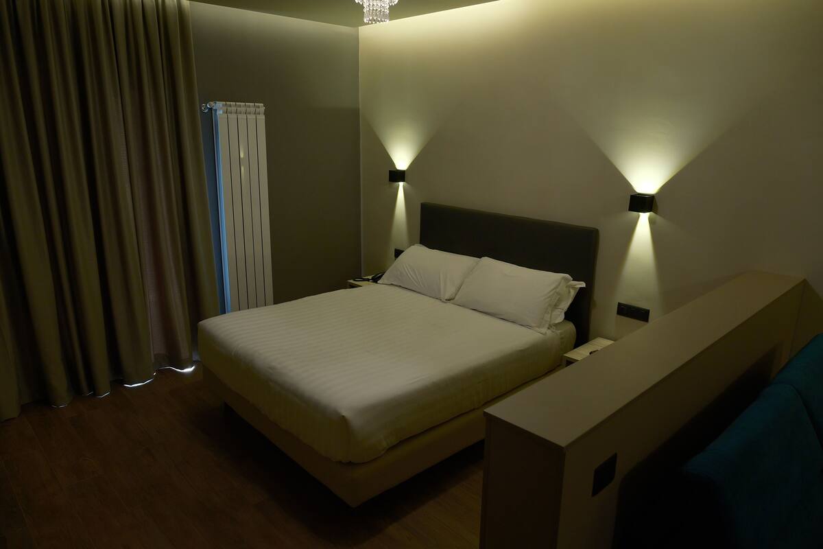 A hotel room with a bed, two bedside tables, a nightstand, a window with curtains, and a ceiling light fixture.
