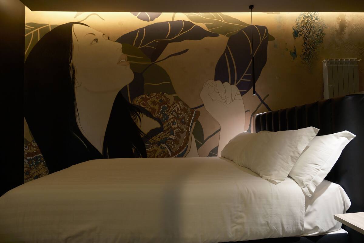 A bed with white linens and a large wall mural of two figures.