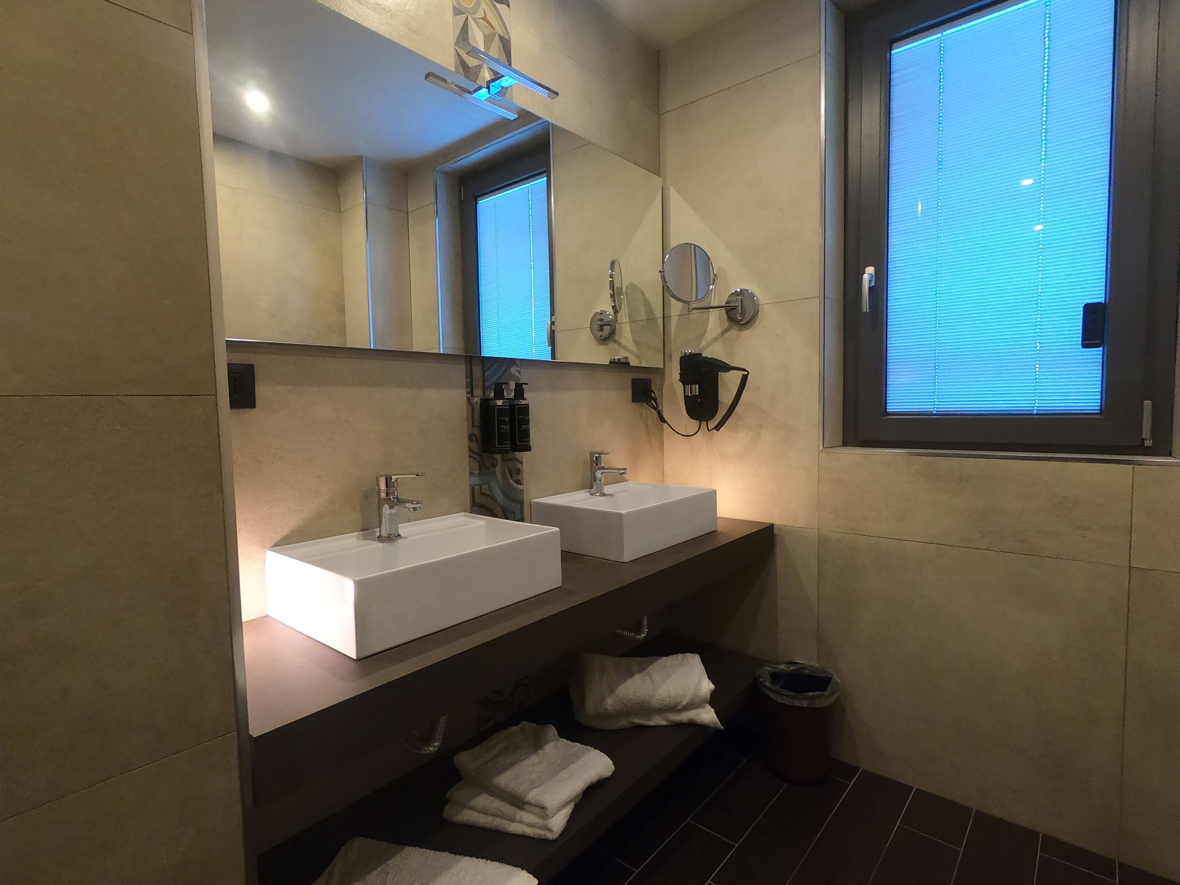 Un baño moderno con dos lavamanos blancos, un espejo grande y una ventana con persianas.