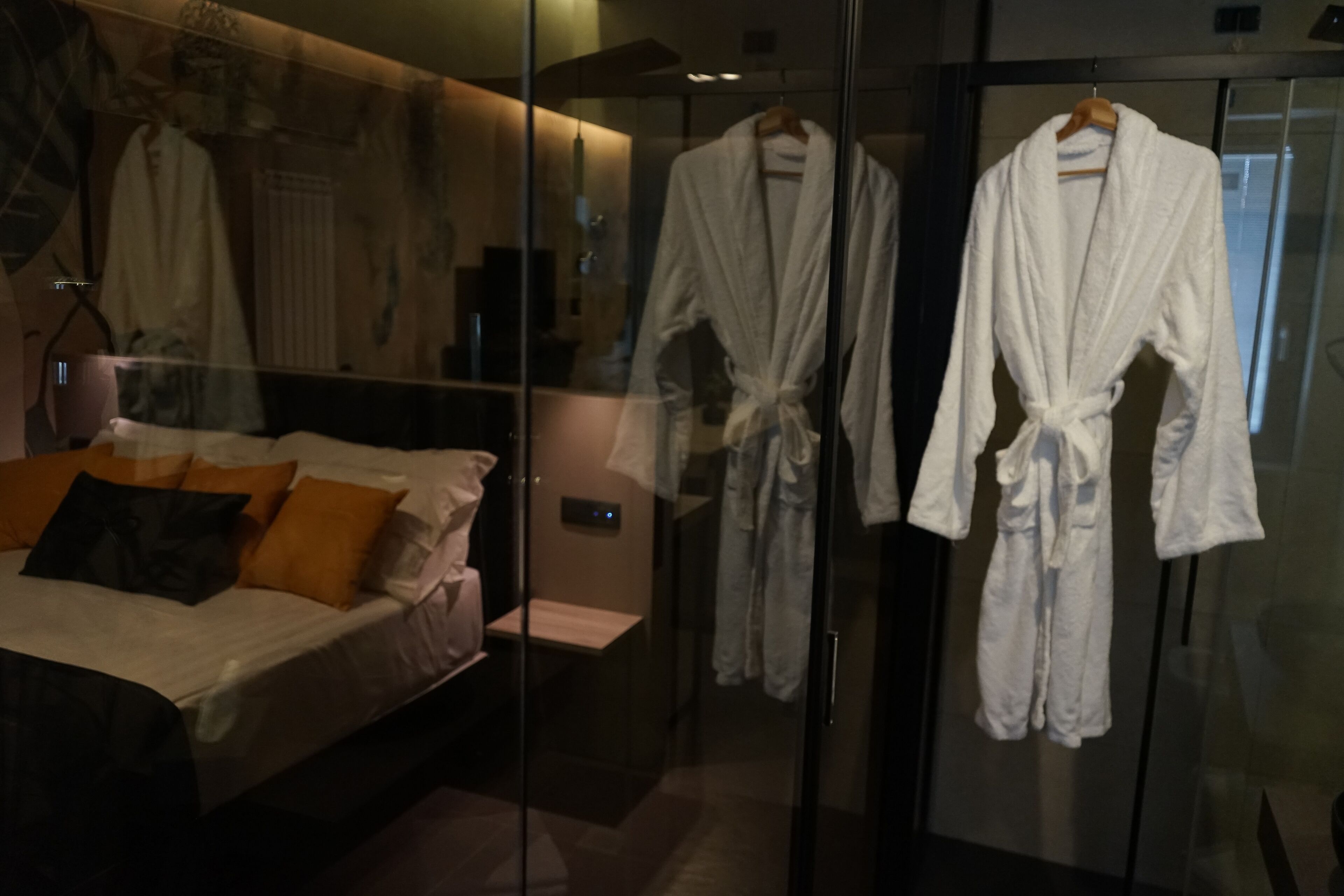 A hotel room with a bed, a glass shower enclosure, and two white robes hanging on a rack.