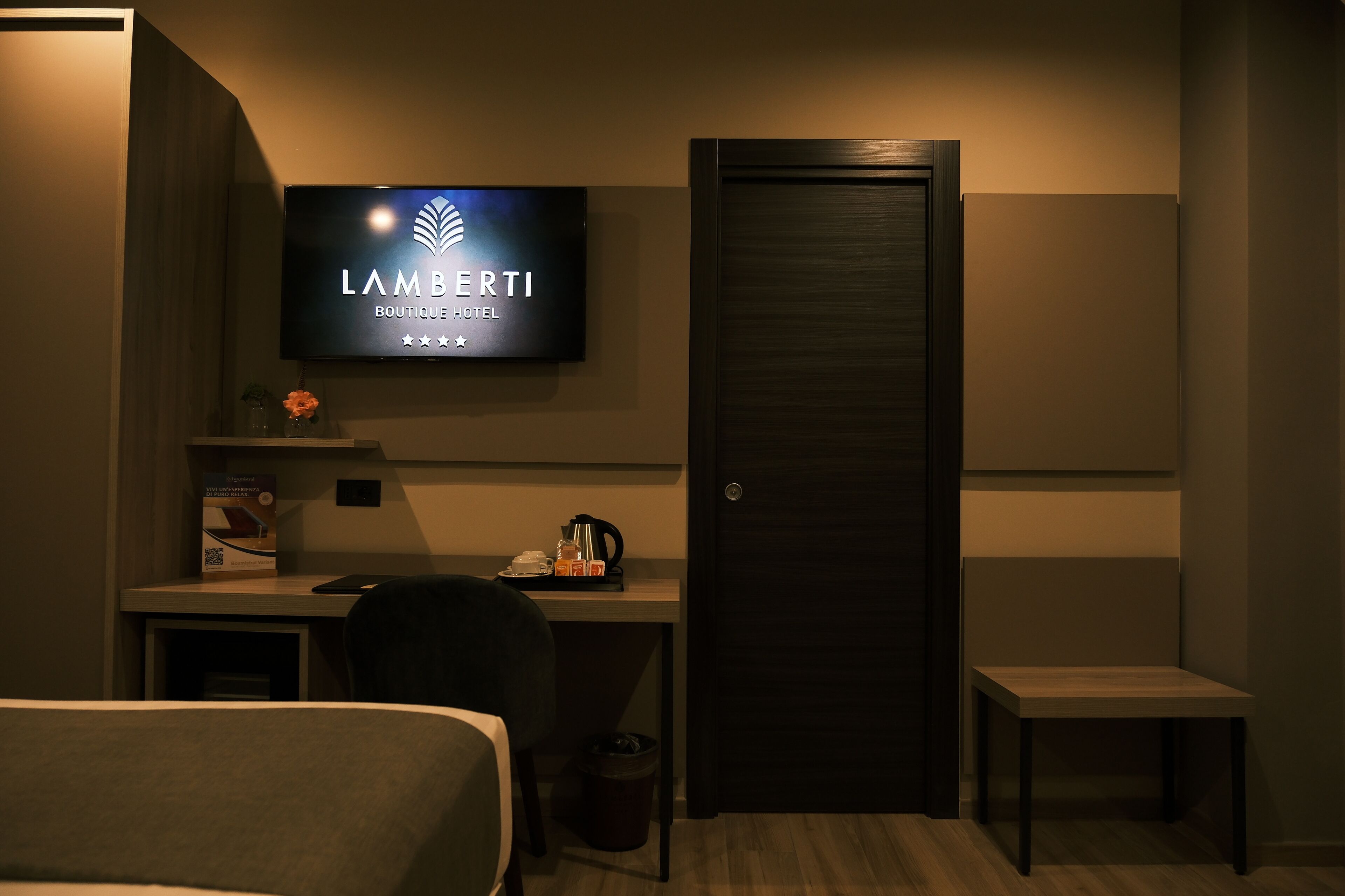 A hotel room with a bed, a desk with a computer, a chair, a small table, a television displaying the hotel name, and a door.