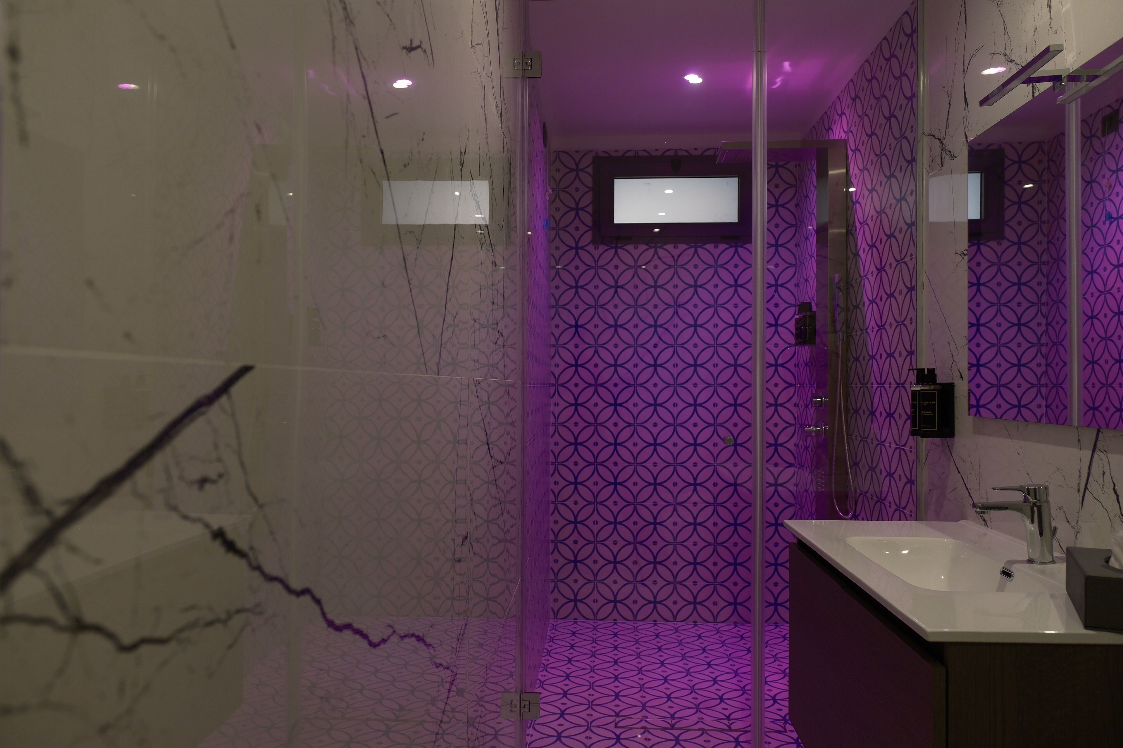 Une salle de bain moderne avec un mur en marbre, un mur orné d’un motif violet, un grand miroir et un lavabo.