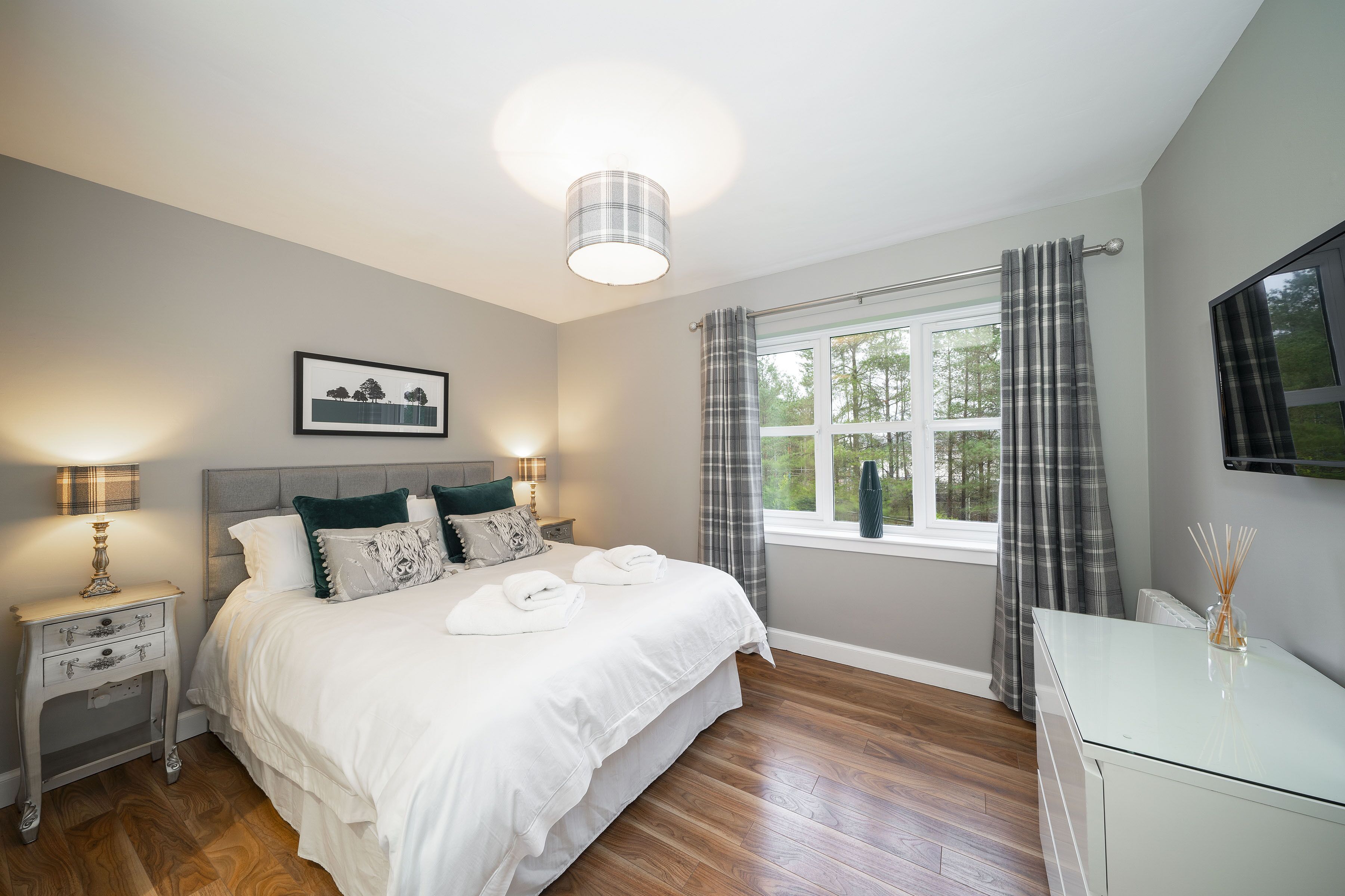 A bedroom with a bed, bedside tables, a lamp, a window with curtains, and a TV on the wall.