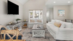 A spacious living room with a ceiling fan, a flat-screen TV, white sofas, a coffee table, and a large window.
