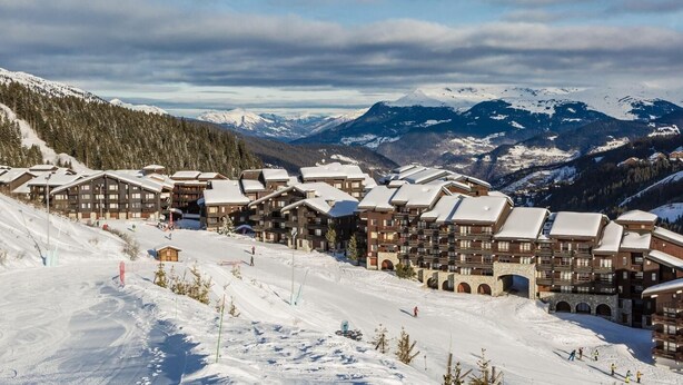 A snow-covered mountain resort with multiple chalet-style buildings, ski slopes, and a panoramic view of snow-capped mountains.