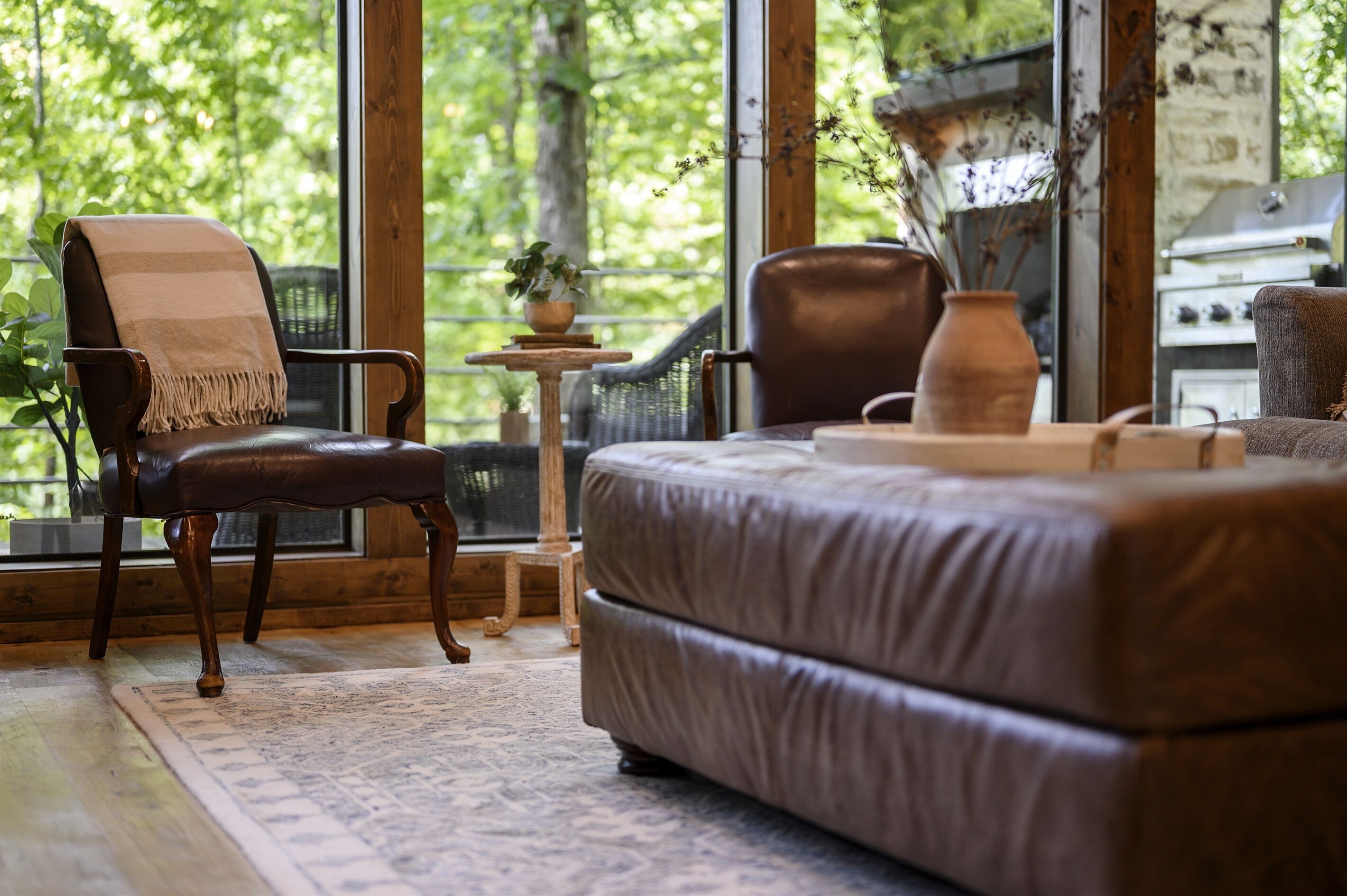 Une pièce chaleureuse avec une grande fenêtre, un canapé en cuir, un fauteuil avec une couverture, une petite table avec une plante et un mur en pierre rustique.
