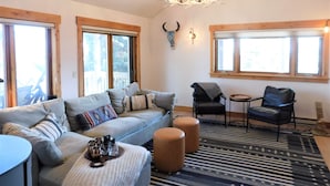 En rummelig stue med en grå sofa, en sort lænestol og et rundt sofabord. Der er en lysekrone i loftet og en oksekraniudsmykning på væggen.