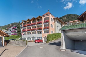 Un hotel de varios pisos con balcones, un estacionamiento y el paisaje de montañas al fondo.