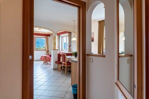 Un couloir mène à une salle à manger équipée d’une table et de chaises, d’une fenêtre avec des rideaux et offrant une vue sur l’extérieur.