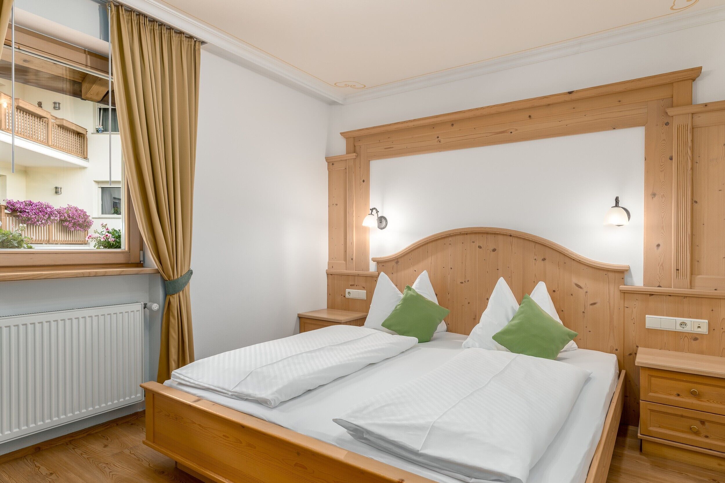 Ein Schlafzimmer mit Holz-Kopfteil, zwei Nachttischen, einem fenster mit Vorhängen und Blick auf einen Balkon mit Blumen.