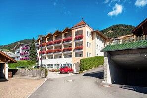 Un hotel de varios pisos con balcones decorados con flores, un estacionamiento y montañas al fondo.