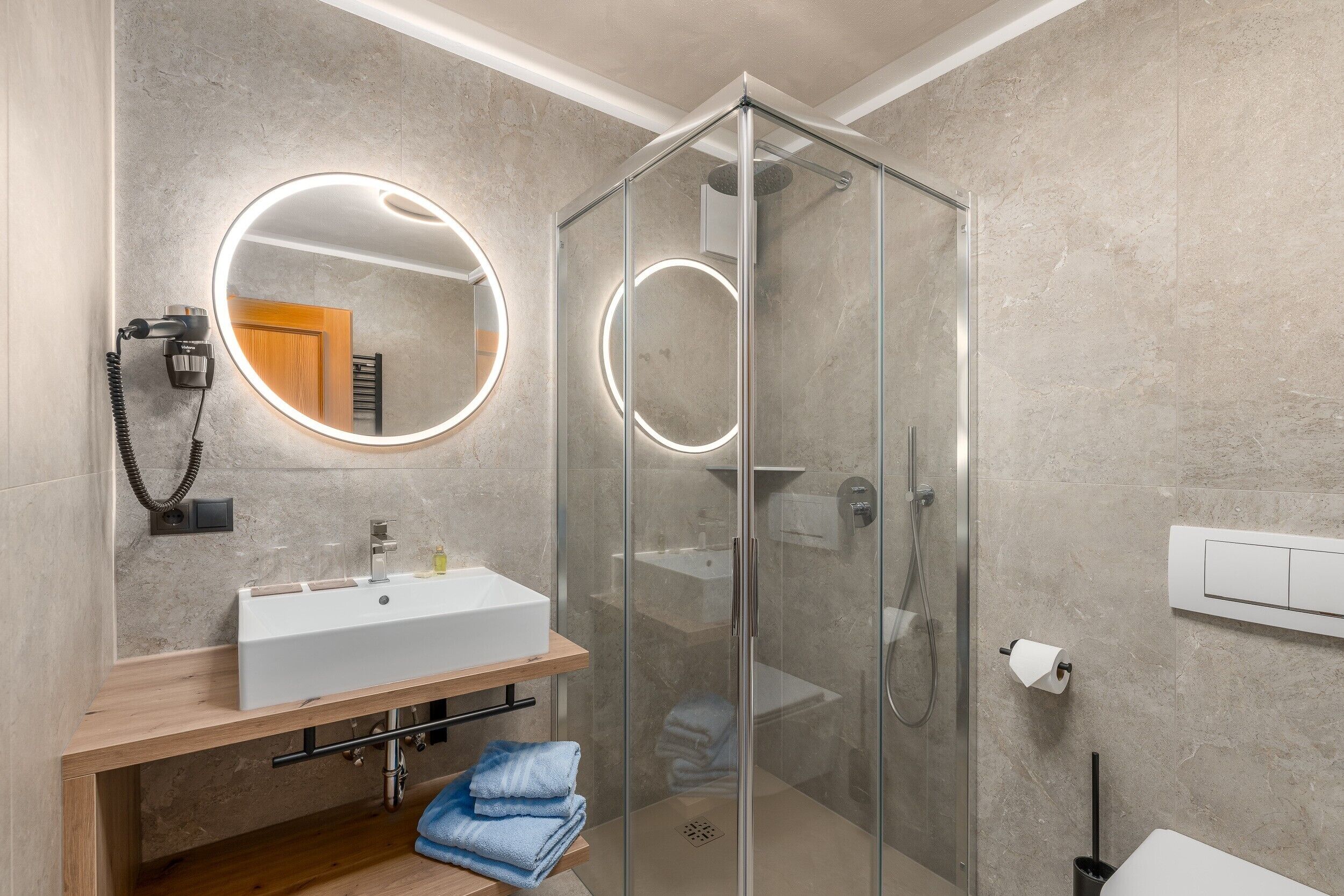 A modern bathroom with a glass-enclosed shower, a round mirror with lights, a wooden vanity, and a towel rack.