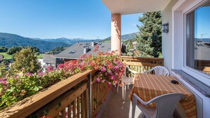 Un balcón con una mesa y sillas, baranda de madera y vistas a montañas y casas.