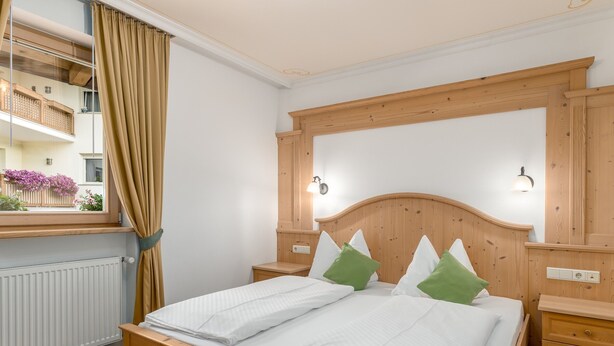 Ein Schlafzimmer mit Holz-Kopfteil, zwei Nachttischen, einem fenster mit Vorhängen und Blick auf einen Balkon mit Blumen.