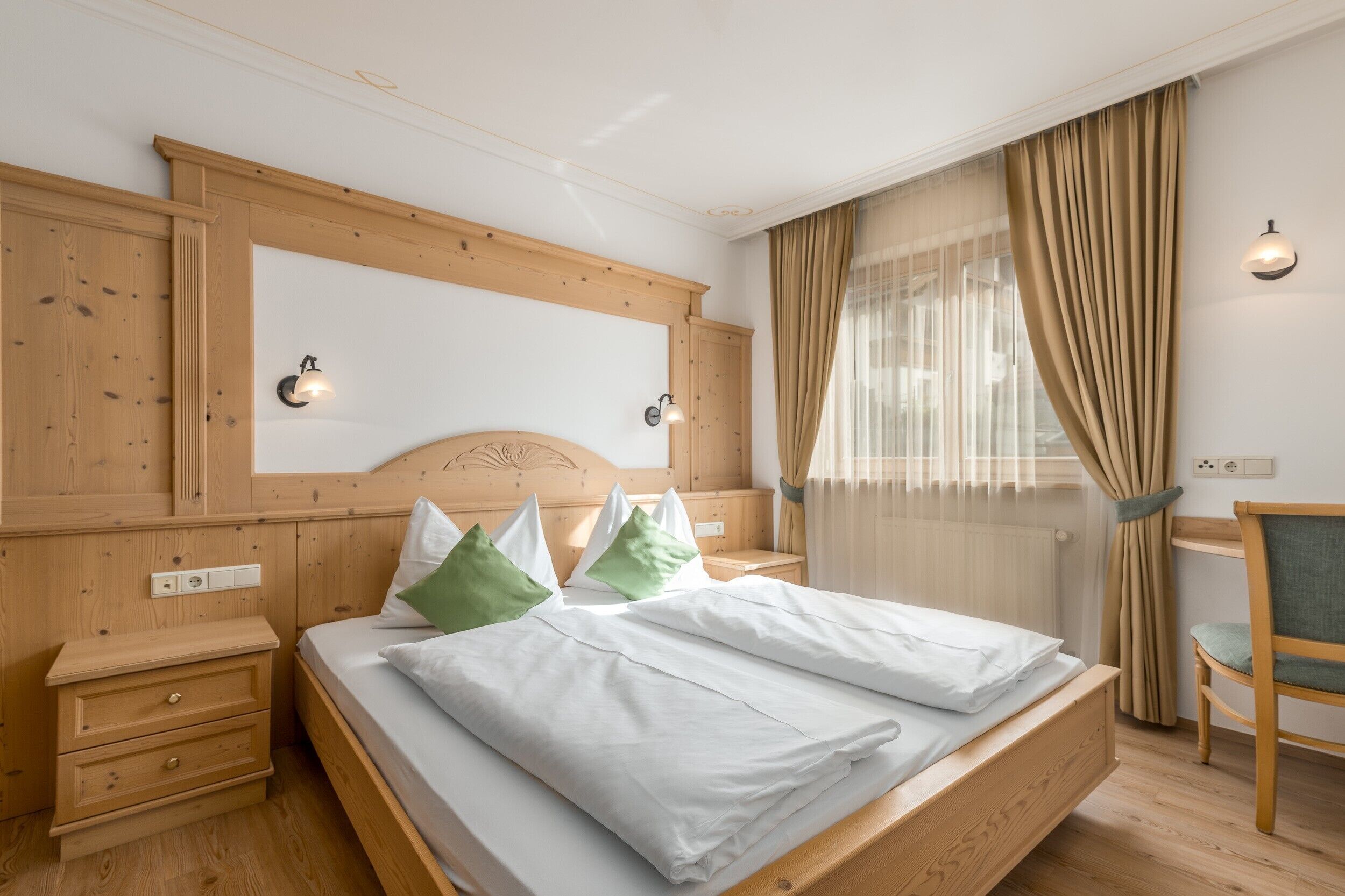 A bedroom with a wooden headboard, a bed with white bedding, a nightstand, a chair, and a window with curtains.