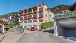 Un hotel de varios pisos con balcones, un estacionamiento y el paisaje de montañas al fondo.