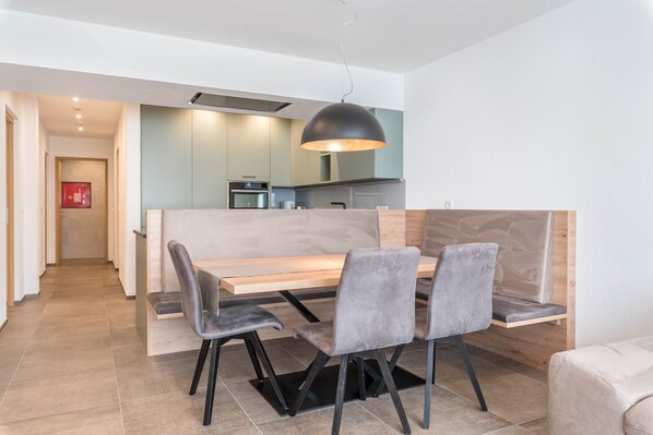 Un espace repas moderne avec une table en bois et des chaises à revêtement gris, un luminaire suspendu et une cuisine en arrière-plan.