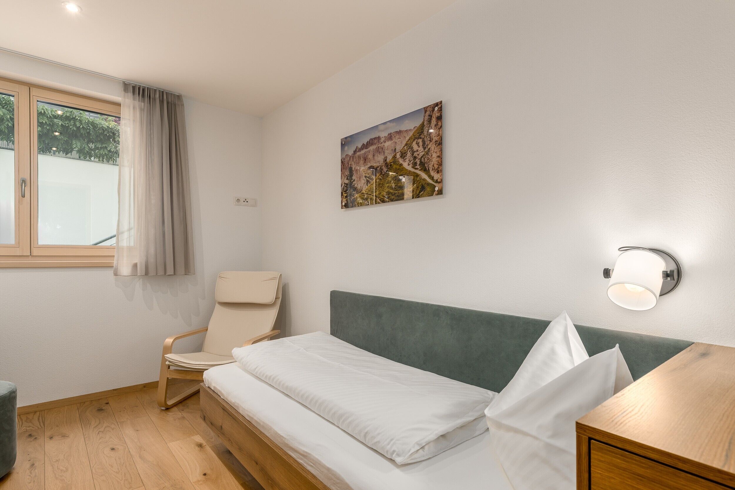 A bedroom with a bed, a chair, a nightstand, a wall-mounted lamp, and a window with curtains.