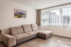 Un salon moderne avec un canapé d’angle, une grande fenêtre ornée de rideaux légers et un tableau représentant un paysage accroché au mur.